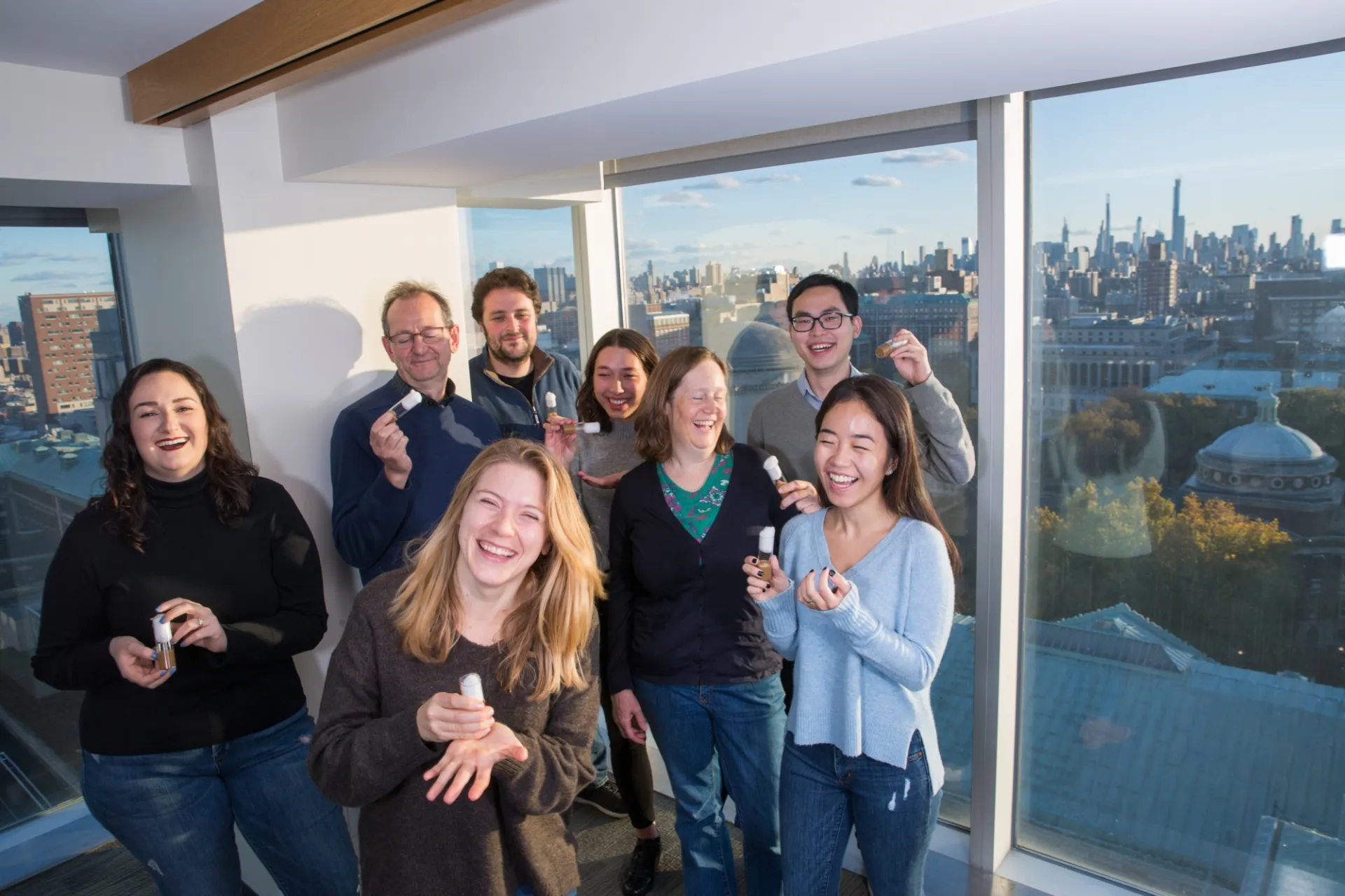 Lab members laughing together and holding fly vials beside windows overlooking campus and the city skyline.